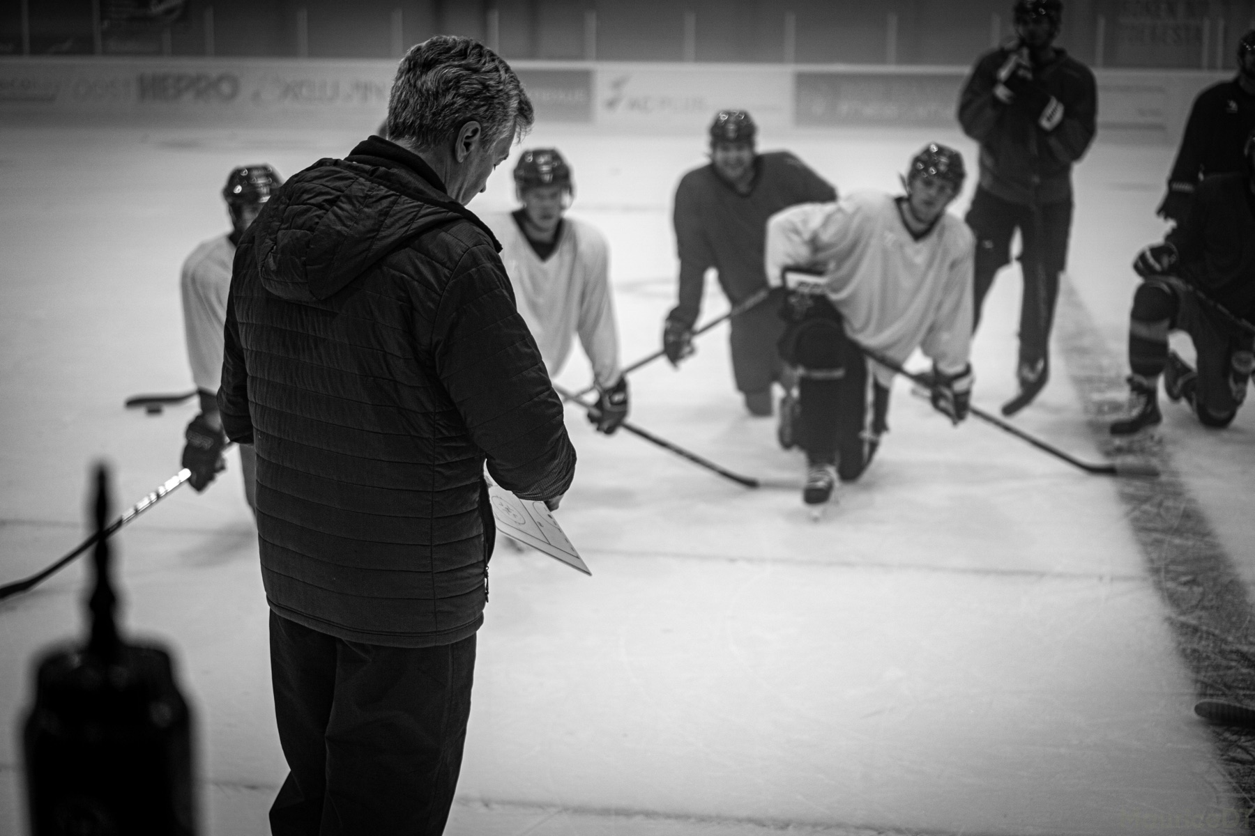A coach reviews a practice plan with hockey players gathered on an indoor ice rink.